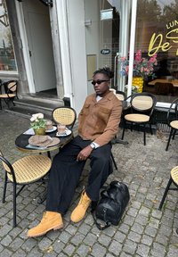 Giacca in suede marrone con due tasche sul petto, pantaloni neri, stivali in suede gialli e una borsa nera. Ambientazione da caffè con un tavolo piccolo e fiori.