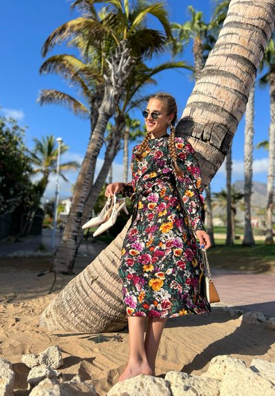 Vestido midi de estampado floral en negro con vibrantes patrones en rosa, amarillo y verde; combinado con zapatos de tacón blancos, de pie cerca de una palmera.
