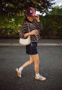 Felpa a strisce bianco e nera, gonna di jeans, cappellino bordeaux, sneakers bianche con dettagli bordeaux e piccola borsa a tracolla bianca, camminando in modo informale.