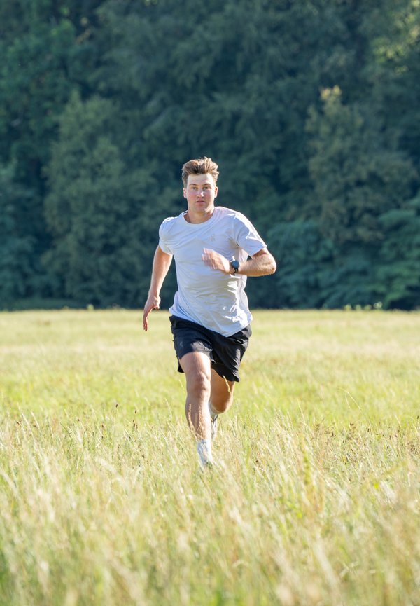 Jeune homme portant une chemise blanche et un short noir courant à travers un champ herbeux avec des arbres en arrière-plan.