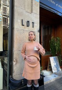 Pruhovaný, pastelově oranžový dvoudílný outfit s cropped topem a sukní. Modelka drží hrnek, stojí před vývěsním štítem, s pozadím ze kamenné zdi.