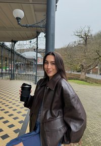 Femme en veste en cuir marron foncé tenant une tasse à café réutilisable, assise sous une galerie ornée avec un sol carrelé et des arbres en arrière-plan.