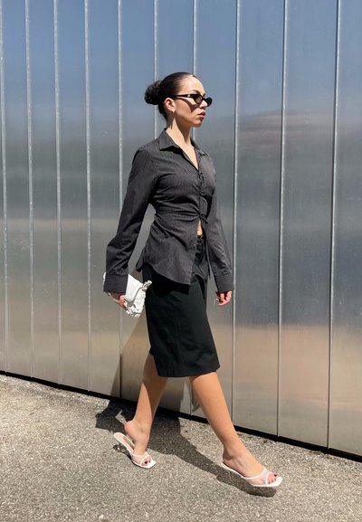 Camisa de botones a rayas blancas y negras, falda negra hasta la rodilla, tacones abiertos blancos, bolso pequeño blanco, caminando contra un fondo metálico.