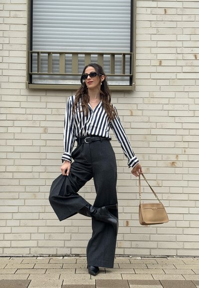 Mujer con gafas de sol negras, camisa de rayas, pantalones de pierna ancha y botas negras posando contra una pared de ladrillo claro con un bolso beige.