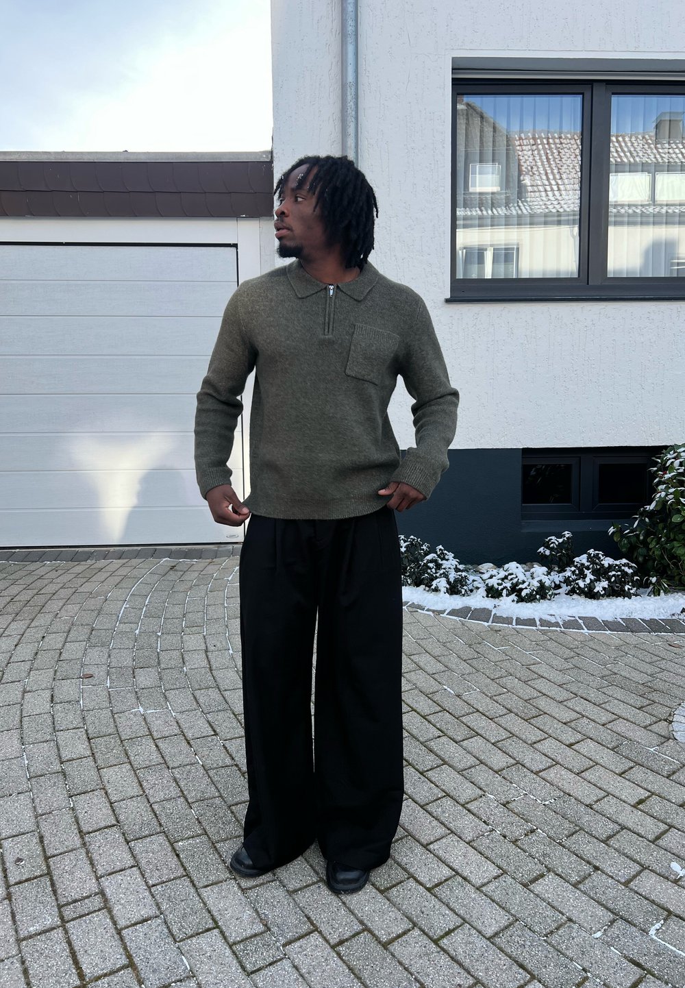 Homme aux dreadlocks portant un pull vert olive et un pantalon large noir, debout sur une allée pavée près d'un parterre de jardin enneigé.