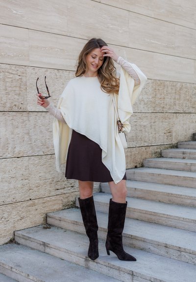 Mujer con poncho crema, falda negra y botas negras hasta la rodilla, de pie en escalones de piedra, sosteniendo gafas de sol y tocándose el cabello.