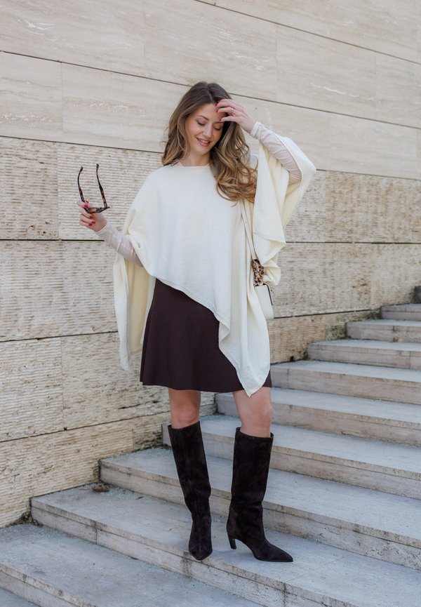 Žena v krémovom ponču, čiernej sukni a čiernych čižmách po kolená stojí na kamenných schodoch, drží slnečné okuliare a dotýka sa vlasov.