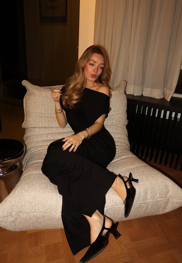 Donna con lunghi capelli mossi che indossa un abito nero con spalle scoperte e tacchi neri, seduta su una poltrona imbottita beige con texture all'interno.