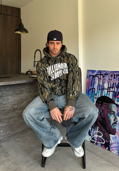 Chaqueta con capucha en estampado de camuflaje con logo blanco, combinada con jeans de mezclilla azul holgados y zapatillas blancas, sentado en un taburete negro.