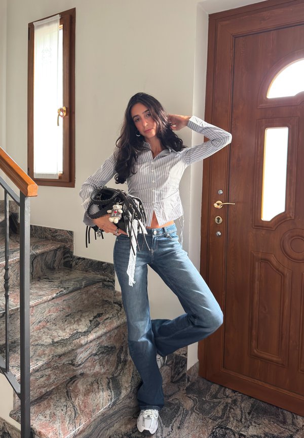 Woman wearing a striped shirt and jeans standing on marble stairs beside a wooden door, holding a black handbag with a keychain, posing with one hand on her head.