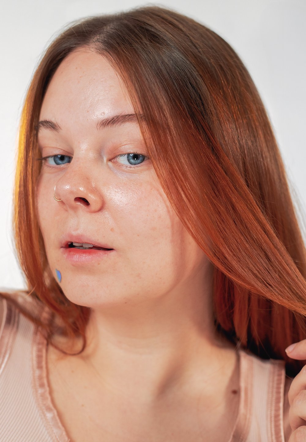 Jeune femme à la peau claire, aux yeux bleus et aux longs cheveux roux tenant une mèche, portant un haut beige et un anneau de nez à la narine gauche.
