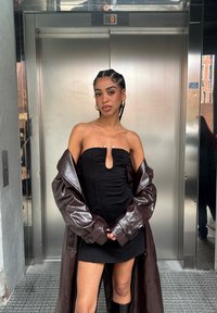 Svart axelbandslös klänning med ett nyckelhålsdetalj, lager-på-lager under en blank brun oversized kappa. Outfiten kompletteras av djärva guldfärgade örhängen.