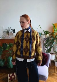 Γυναίκα με κόκκινα μαλλιά, που φοράει καφέ και κίτρινο πουλόβερ με σχέδιο αργκολ και μπλε φουλάρι με πουά, στέκεται σε δωμάτιο με φυτά και έπιπλα.