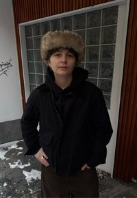 Veste noire à capuche avec de grandes poches, portée sur un sweat à capuche noir. Assortie à une jupe en velours côtelé marron. Chapeau doublé de fourrure ajoutant de la texture. Sol enneigé visible.