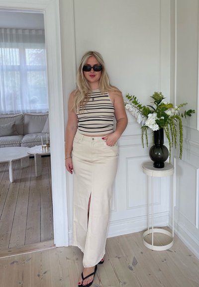 Top sin tirantes con rayas negras, falda maxi color crema con una abertura frontal, chanclas negras y gafas de sol, todo en una habitación iluminada.