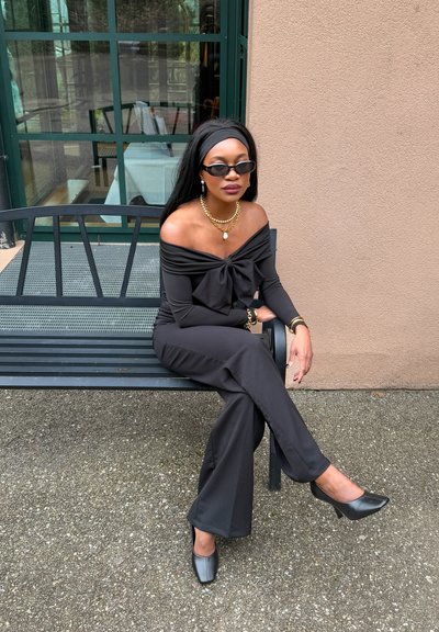 Monos de color negro con los hombros al descubierto, con un detalle de lazo, combinado con tacones negros. Los accesorios incluyen un collar dorado y gafas de sol. Sentada en un banco.