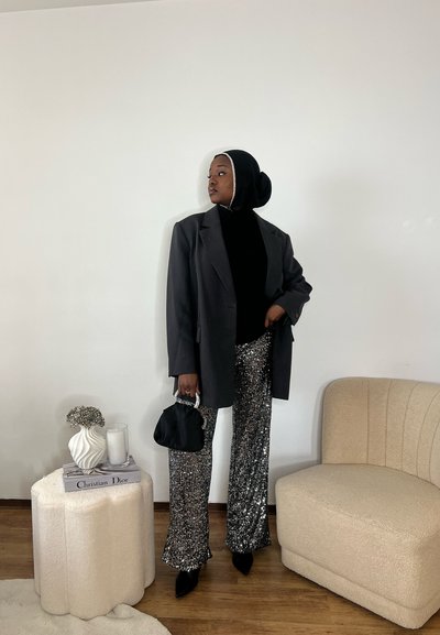 Blazer negro oversize, cuello alto negro y pantalones acampanados con lentejuelas metálicas. Bolso negro con asa. Silla texturizada color crema. Fondo de pared blanca.