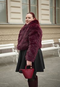 Γυναίκα με μπορντό γούνινο μπουφάν, τσάντα και μπότες που ταιριάζουν, μαύρη φούστα, στέκεται σε πεζοδρόμιο πόλης κοντά σε λευκά παγκάκια.