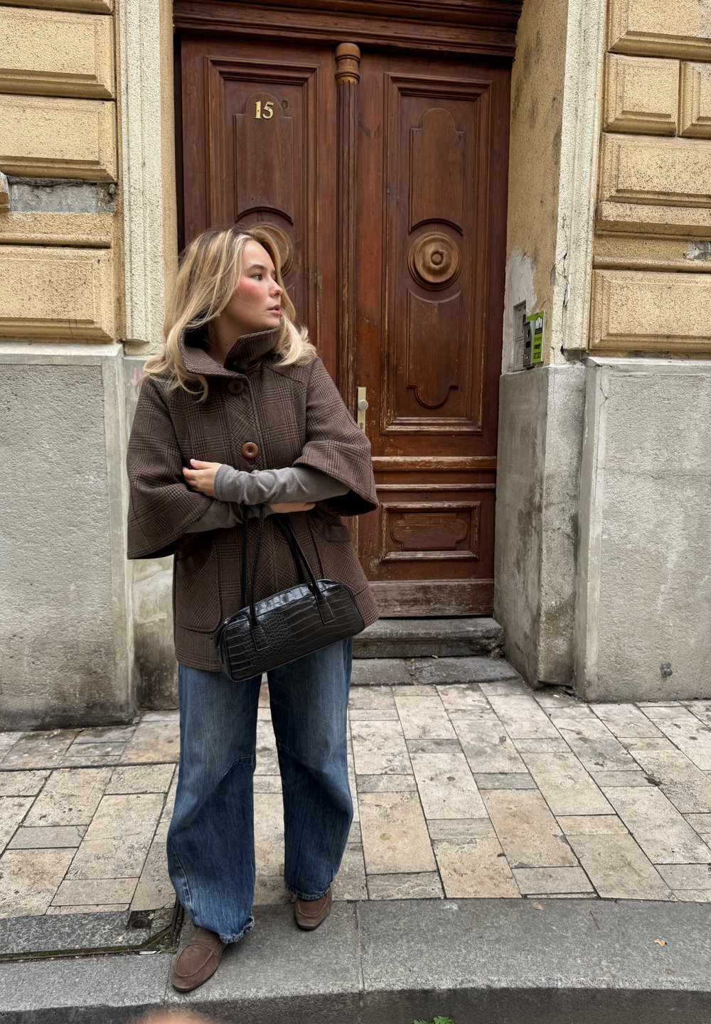 Abrigo grande de color marrón y texturizado con un cuello alto, dos botones, combinado con mangas largas grises, vaqueros anchos azules y un pequeño bolso negro con relieve de cocodrilo.