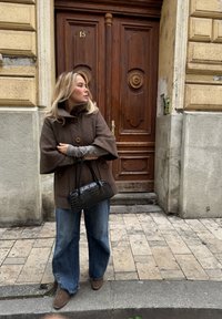 Abrigo grande de color marrón y texturizado con un cuello alto, dos botones, combinado con mangas largas grises, vaqueros anchos azules y un pequeño bolso negro con relieve de cocodrilo.