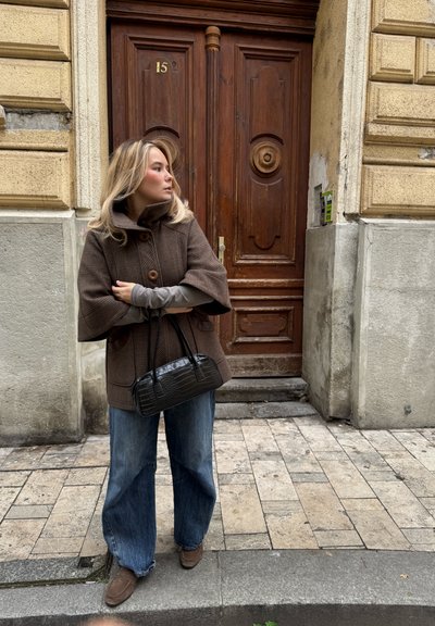 Abrigo grande de color marrón y texturizado con un cuello alto, dos botones, combinado con mangas largas grises, vaqueros anchos azules y un pequeño bolso negro con relieve de cocodrilo.