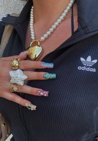 Sort zip-up jakke med hvid Adidas-logo; stor perlekæde; hænder, der viser farverige, udsmykkede akrylnails og forskellige ringe.