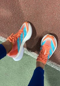 Persoon met feloranje en blauwe hardloopschoenen die op een rood-groene buitenbaan staat, met voeten die een witte lijn kruisen.