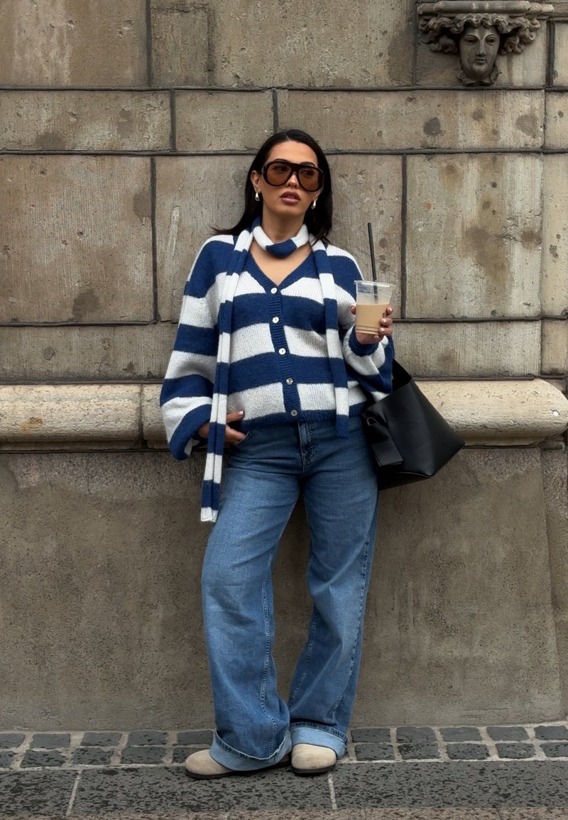 Cardigan rayé bleu et blanc, jeans bleus, chaussures beiges et un sac fourre-tout noir. Tenant une tasse transparente avec une paille.