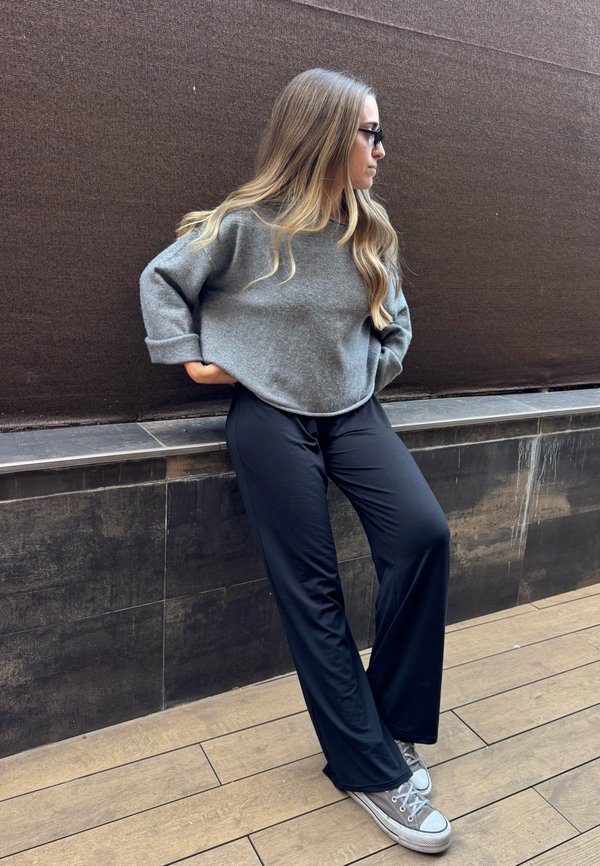 Une femme portant des lunettes, un pull gris, un pantalon bleu marine et des baskets s’appuie contre un mur carrelé.