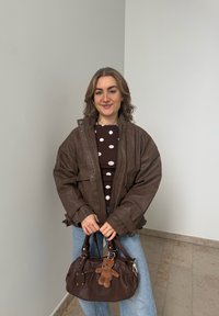 Jeune femme portant une veste en cuir marron et une chemise à pois, tenant un sac à main marron avec un porte-clés ourson, debout devant un mur uni.