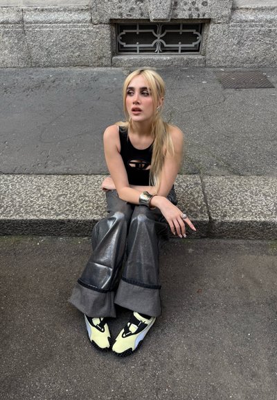 Top corto negro con detalle de corte, pantalones anchos metálicos plateados, zapatillas amarillas con acentos negros, sentada en una acera de piedra.