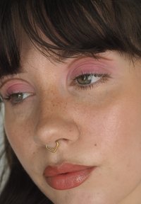 Nærbilde av et ansikt med rosa øyenskygge, gullhjerteformet nesering og glinsende nude lepper. Huden har fregner og en jevn tekstur.