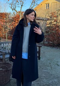 Manteau en laine noire avec capuche, pull gris clair en dessous, superposé sur une chemise bleue. Texture lisse, longueur genoux, fermeture à boutons sur le devant.