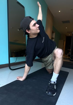 Una camiseta negra y pantalones cortos caqui en una persona que realiza un ejercicio sobre una esterilla negra, con una pared azul aqua y un gran espejo de fondo.