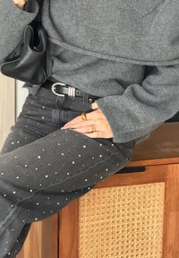 Kvinne iført grå genser og svarte jeans med sølvnitter, svart belte, holder svart skinnveske, sitter på et treskap med flettet mønster.