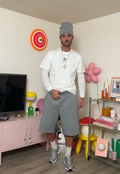 Un joven con gorro gris, camisetas blancas superpuestas, pantalones cortos grises y zapatillas de deporte está de pie en una habitación colorida con una televisión, un armario rosa y objetos decorativos.