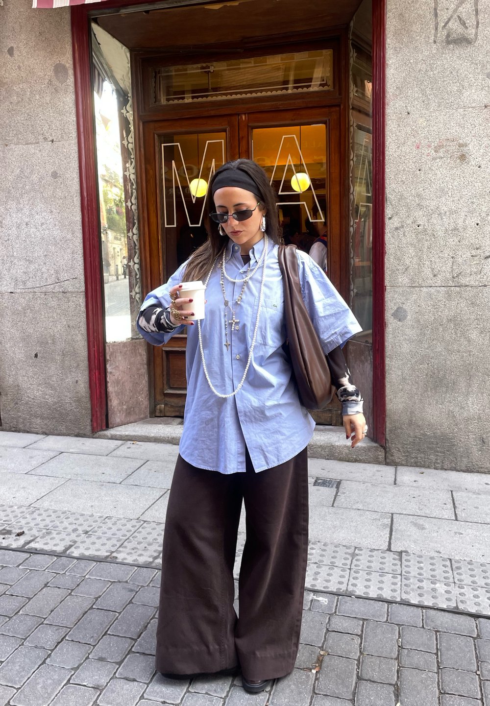 Light blue oversized shirt, brown wide-legged trousers, brown shoulder bag, layered necklaces, and black sunglasses, standing on a cobbled street.
