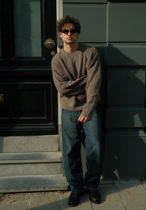 Joven con pelo rizado y gafas de sol, vestido con un suéter marrón y vaqueros azules, de pie junto a la entrada oscura de un edificio bajo la luz del sol.