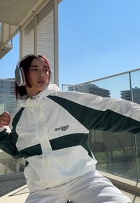 Giacca a vento bianca e verde con chiusura a zip, pannelli diagonali a contrasto e un piccolo logo, indossata con cuffie argento.