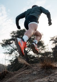 Chaussures de course avec des détails orange et des accents noirs, semelle extérieure texturée et bandes blanches. Du sable est déplacé alors qu'une personne saute.