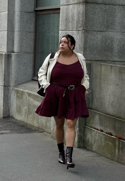 Una mujer con un vestido burdeos en capas y cinturón en la cintura, combinando con una chaqueta blanca y brillantes botines negros, camina por una acera.