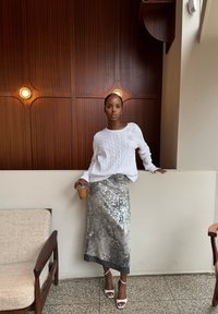 Vitt kabelstickat tröja kombinerad med en silverfärgad paljettskjort. Håller en genomskinlig kopp, står mot en träbakgrund, bär vita klacksandaler.