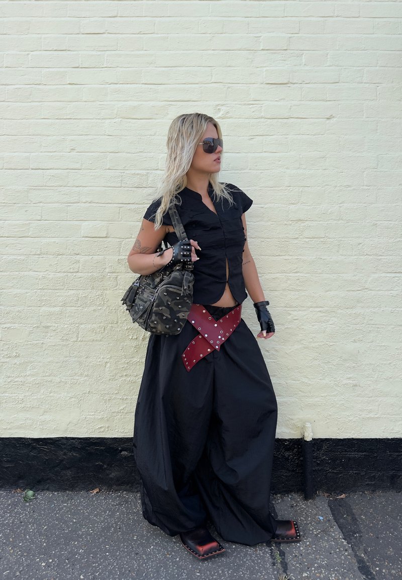 Femme aux cheveux blonds portant des lunettes de soleil, un haut court noir, un pantalon large noir avec une ceinture rouge, des gants sans doigts, et portant un sac camouflage.