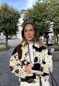 Kurtka z futerka ekologicznego z patchworkiem czarnych i brązowych plam na kremowym tle, z zamkiem błyskawicznym i kołnierzem. Noszona na białej koszuli.