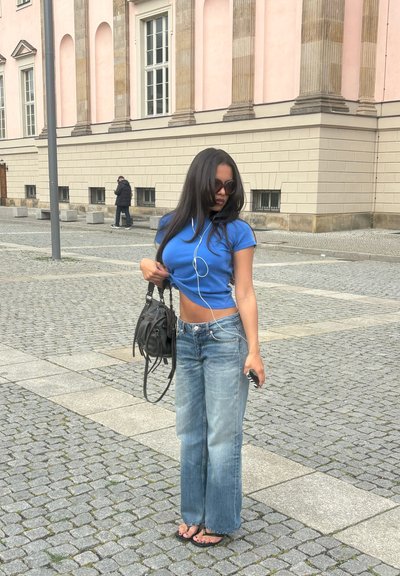 Joven mujer que lleva un top corto azul, vaqueros, gafas de sol y sandalias, está de pie en una calle adoquinada cerca de un edificio beige, sosteniendo una bolsa negra.