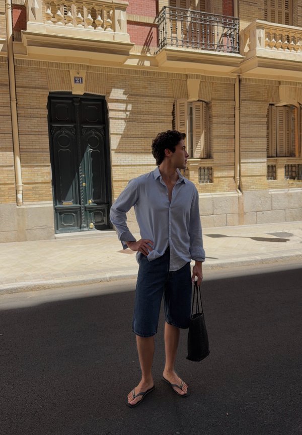 Jeune homme en chemise décontractée, short et tongs, tenant un sac fourre-tout, debout dans une rue avec des façades de bâtiments ornées en arrière-plan.