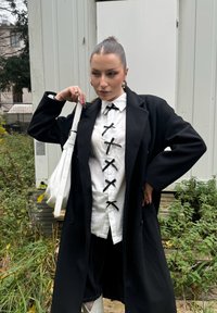 Svart långkappa över en vit skjorta med svarta band, parat med svarta shorts och en vit handväska. Silverörhängen i båge och ringar.