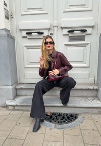 Maronfarvet læderjakke, sort top, sorte brede jeans og støvler med spidse tæer. Solbriller fuldender outfittet. Sten trin og dekorativ dræn dæksel.