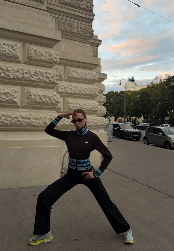 Una persona con gafas de sol y ropa negra con rayas azules posa junto a un edificio de piedra ornamentado en una acera de la ciudad.