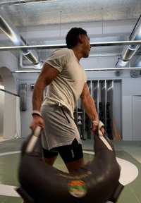 Homme balançant un sac de sable lourd dans une salle de sport moderne avec des tuyaux industriels au plafond et des poids sur des supports en arrière-plan.